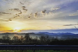 Over a metal railing, a pale yellow Appalachian sun rises over the Blue ridge Mountains, seen in the distance. Its cold, and the fog is thick. The stratus clouds are scarce, and feathered. It makes me sad.