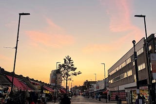 A picture of a market road in London in the evening with few people walking around, with a beautiful summer sunset in the background