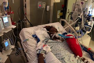 Image of the author, unconscious in a hospital bed, intubated and connected to many medical devices with a heart surgery pillow by her side.