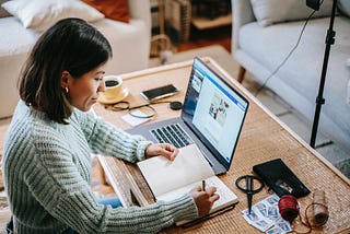 Woman writing in a notebook next to her laptop.