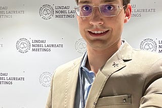 Selfie de Gustavo Rosa Gameiro, ele é um homem branco jovem, usa óculos, paletó beje e camisa azul e ao fundo está uma parede com o marca e texto do “Lindau Nobel Laureate Meetings”