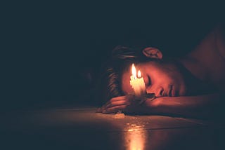 A man with well-style hair and minimal facial hair lays on his side, one arm in front of him. On his wrist, just in front of his face, are several melted together candles, lit, providing the only light in the image. The man’s eves are closed, almost as if he is asleep, wax dripping down the candles onto his wrist and the floor under him.