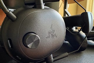 A Razer BlackShark V2 HyperSpeed headset lying on its side on a desk mat against a controller, with the Razer logo and large volume knob highlighted in the center of the frame.
