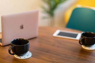 A table with a laptop, an ipad, and two coffee cups sitting on it.