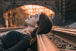 A woman reclines with her head on a railway track, railway bridge in the background