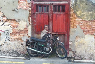 Old brick wall and reddish brown wooden door, in front of it is a motorcycle leaning against the door, a painting of a boy appears to be sitting on the motorcycle and looking over his shoulder at a line drawing of a dinosaur