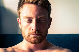 Head shot, shoulders up, of bare chested red-headed man with his eyes closed in meditation.