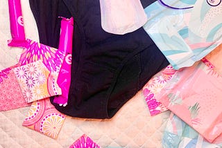 A variety of blue and pink floral print pads, tampons and dark coloured period panties randomly sprawled out across a white background