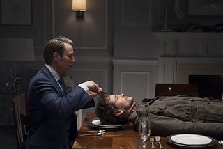 Dr Hannibal Lecter sits in a suit at a dinner table with a man (Will Graham) lying on his plate. Lecter has a knife and fork in his hands as if he’s about to eat Graham.