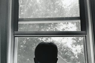 Black and white photograph of the back of a boy in front of a window.