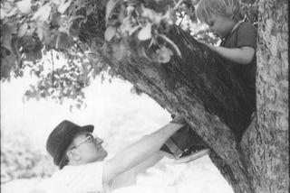 Little boy sitting up in a tree. He is being lovingly held in place by his grandfather.