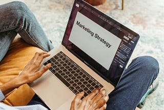 Laptop with screen reading “Marketing Strategy” on the lap of a man wearing blue jeans. Kind of on the nose. Sorry.