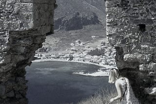 A young woman wearing a summer dress peers through a ruined stone arch at a coastal village far below.