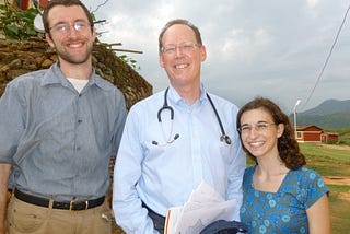 Paul Farmer stands between two GlobeMed students. He is wearing a long-sleeve, light blue button up shirt with a stethoscope slung around his neck. To the left is a student who is taller than Paul, has some scruffy facial hair, and is wearing a short-sleeve, gray button up. To the right is a student who is shorter than Paul and is wearing a blue patterned top with a square neckline. They are all smiling at the camera, with a rural landscape in the background.