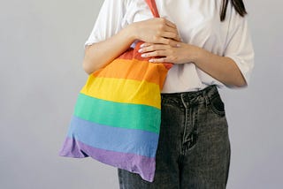 Person in grey jeans and white tee-shirt carrying a rainbow totebag.