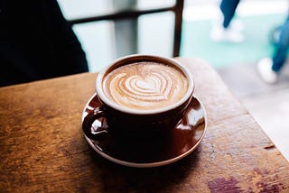 A cup of coffee on a worn out wooden table. The coffee has a heart drawn in milk, and is in a red mug on a matching saucer.