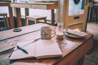 A wooden table with an open journal, mug, glass of milk, and a small plate on top.