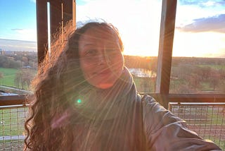 Woman with long, curly hair is smiling while looking into the camera with the sun setting behind her. The rays from the sun are shining across her face. The sky and grass are in the background.