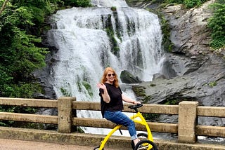 White woman with red hair sits on an yellow walking bike on a bridge in front of a water fall.