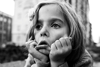 A black-and-white image of a light-skinned young girl. A close-up on her face set against a blurry background of old-brick apartment buildings. She has her head resting in her hands and she’s pulling her cheeks down, forming her face into a exagerrated frown.