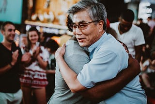 A man with a smile, hugging another, other people are in the background, smiling and a clapping.