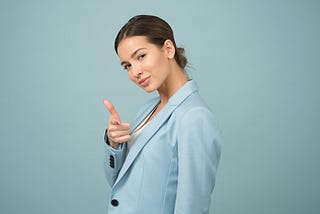 woman in blue coat, dark hair, pointing at camera, blue background