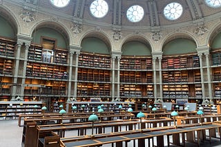 Vast BNF Richelieu library with vaulted ceiling, ornate arches and three levels of shelves overflowing with books. Reading tables with green lamps are neatly arranged, inviting study and reflection.