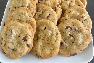 A dozen chocolate chip cookies on a white square plate on a grey table