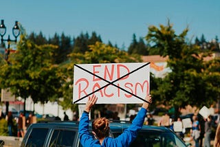 Protester holdiong adn “End Racism” sign at a rally. The author has used Microsoft Paint to create a big black “X” over the sigm.