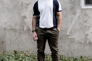 Félix stands outside on a sunny day, in ankle high greenery, in front of a gray wall. He is bald, with round glasses and a beard. He is wearing a navy and white striped shirt and army green pants, with his left hand in his pocket.