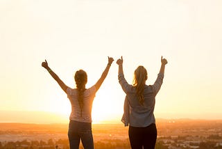 two women standing in front of a bright orange sunset with their arms in the air