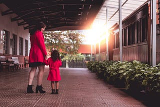 A woman and a little girl with short brown hair holding hands, dressed in red coats and black boots standing under a patio outdoors.