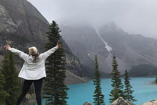 Woman standing on a rock facing mountains in the fog and a bright blue lake with arms spread wide open.