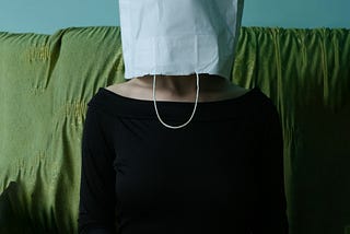 A woman sits on a green sofa, her head covered by a white paper bag. She’s wearing a black top and has her hands in her lap.