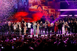 Wide, vibrant shot of a star-studded tribute concert: dozens of artists and celebrities line up on a brightly lit stage under purple and red spotlights as multicolored confetti rains down, performing in front of a packed arena crowd holding up smartphones.