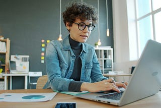Small-business owner works at a laptop in an organized office with charts and notes visible, illustrating workflow management and business systems. Photo by Vitaly Gariev on Unsplash