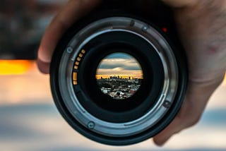 Close up of hand holding a camera lens, with view of distant city in focus.
