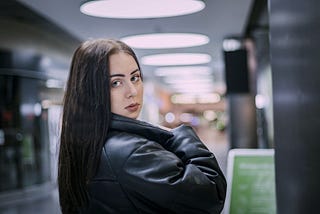 An attractive dark-haired girl in a black jacket is standing in an airport concourse, looking back over her shoulder towards the camera.