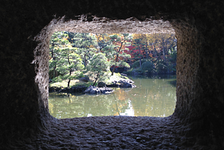 A photo taken through a Japanese stone lantern, looking to a bunch of small green trees on a grassy mound in the middle of a murky, green-brown lake. Additional trees fill the background. It’s a bright, sunny day. The image is framed by the darkness of inside the stone lantern.