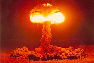 A color photo of a mushroom cloud from an atomic text at Nevada Test Site in 1957. The photo is very orange with the top of the mushroom cloud in very bright light.