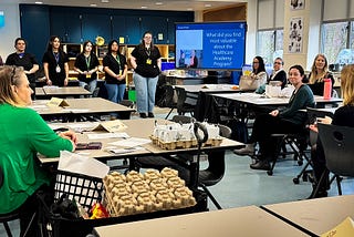 Six high high school students stand in front of a classroom giving a presentation to several adults sitting at desks.