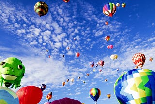 Myriad colorful hot air balloons rising into a blue sky flecked with white, puffy clouds.