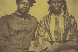 A photograph of young Black couple. The man wears a uniform can cap and looks down. His arm is draped around the shoulders of a pretty young woman in braids, elaborate earrings, and a checked dress.