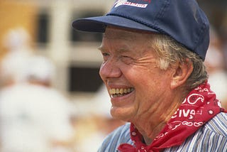 A closeup photo of former President Jimmy Carter, at a work site, wearing a neckerchief and baseball cap.