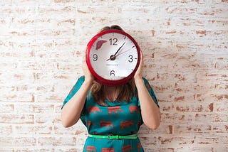 A woman wearing a green dress with red elephants on it holding a red clock in front of her face.