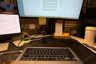 A well-lit desk setup features a MacBook with its lid open, positioned in front of a large curved monitor displaying a Google Docs document in editing mode. A smaller tablet screen is visible to the left. The desk is covered with a black and geometric-patterned mat, with a white keyboard, a mouse, a pen, and sticky notes. A Starbucks cup sits on the right side. Behind the monitor, there are framed photos on the wall, and a small note with handwritten text is attached to the monitor stand.