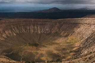 A vast, empty crater with distant mountains, ominous clouds and peeking sunlight in the background