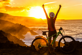 A woman standing in front of her bike, with the helmet, raising her hands up looking at the sunrise