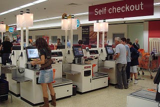 Three self-checkout machines at a store with a woman purchasing something at the first one and a man and son at the 3rd one