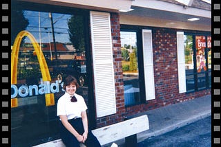 A teenage girl wearing McDonald’s uniform is sitting in front of McDonald’s where she works. She is smiling.